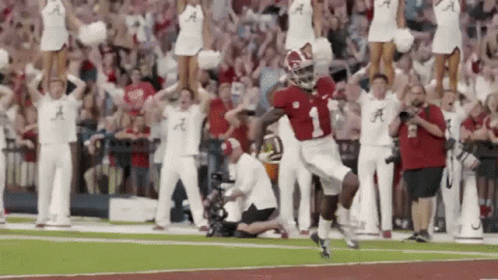 A Football Player Is Running On The Field With Cheerleaders Behind Him And A Crowd Watching . GIF