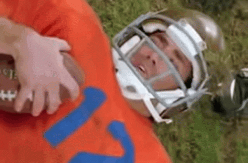 A Football Player Wearing A Helmet Is Laying On The Grass With A Football . GIF