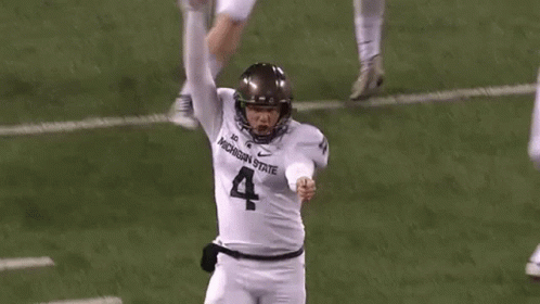 a football player wearing a michigan state jersey is kicking a ball on the field .