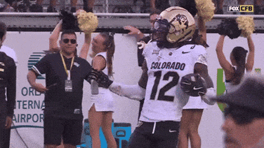 A Football Player With The Number 12 On His Jersey Stands In Front Of Cheerleaders GIF