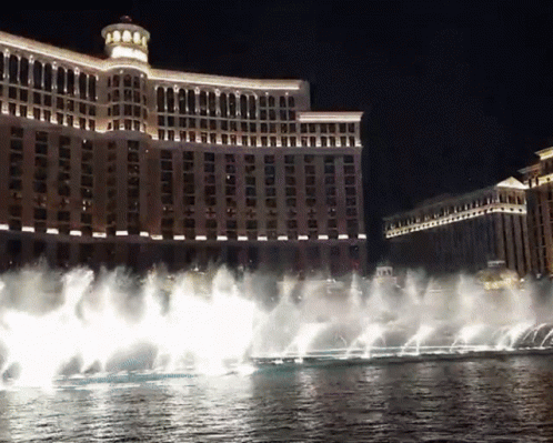 A Fountain In Front Of A Large Building That Is Lit Up At Night GIF