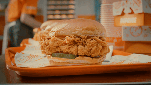 A Fried Chicken Sandwich On An Orange Tray With A Pickle On It GIF