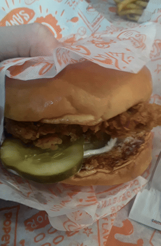 A Fried Chicken Sandwich With A Pickle On Top Is Wrapped In Paper That Says Shakes GIF