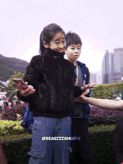 A Girl In A Black Sweater Is Holding A Cell Phone While Standing Next To A Boy In A Blue Jacket GIF