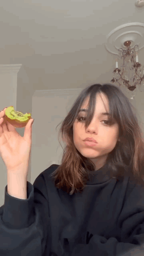 A Girl In A Black Sweatshirt Is Holding A Slice Of Kiwi In Her Hand GIF