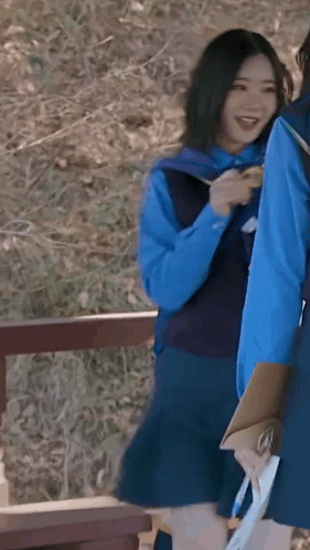 A Girl In A Blue Vest And Skirt Is Holding A Clipboard And Smiling While Standing Next To Another Girl . GIF