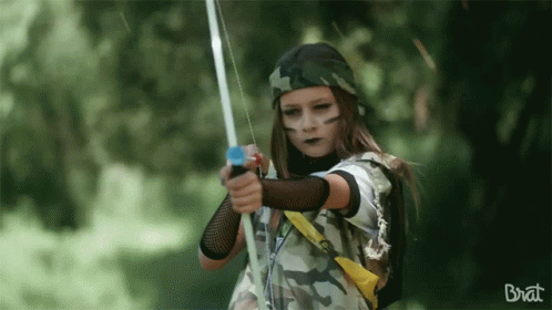 A Girl In A Camouflage Outfit Is Holding A Bow And Arrow And The Word Brat Is On The Bottom GIF