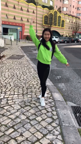 a girl in a neon green sweatshirt and black pants is standing on a cobblestone sidewalk .