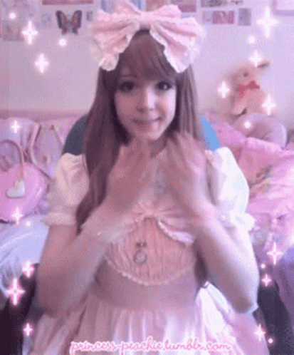 A Girl In A Pink Dress With A Pink Bow On Her Head Is Making A Heart With Her Hands . GIF