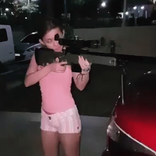 A Girl In A Pink Shirt And White Shorts Is Holding A Gun In Front Of A Red Car . GIF