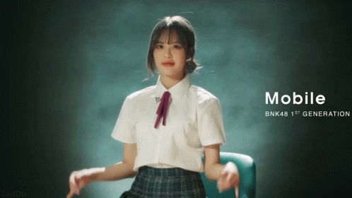 A Girl In A School Uniform Is Sitting In A Chair With The Words Mobile Bnk48 1St Generation On The Bottom GIF