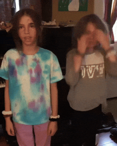 A Girl In A Tie Dye Shirt Is Standing Next To Another Girl Wearing A Sweater That Says Jv GIF