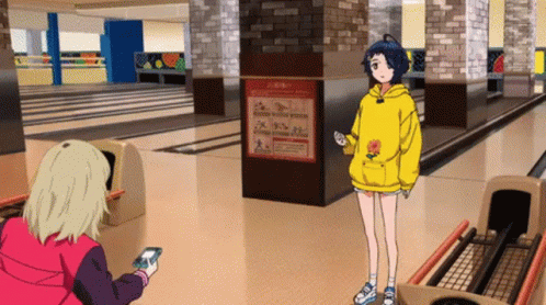 A Girl In A Yellow Hoodie Is Standing In A Bowling Alley While Another Girl Looks At Her Phone . GIF