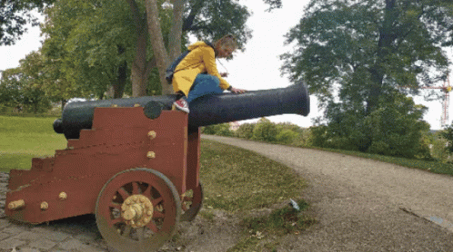A Girl In A Yellow Raincoat Is Sitting On A Cannon GIF