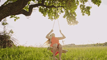 A Girl In An Orange Dress Is Sitting On A Wooden Swing Under A Tree GIF