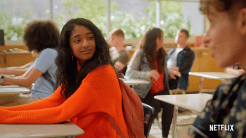 A Girl In An Orange Sweater Sits At A Desk In A Classroom With A Netflix Logo On The Bottom GIF