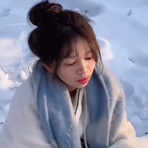 A Girl Is Sitting In The Snow With Her Eyes Closed And A Blue Scarf Around Her Neck GIF
