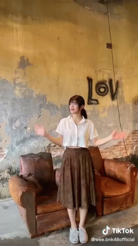 A Girl Is Standing In Front Of A Couch With The Word Love Written On The Wall Behind Her GIF