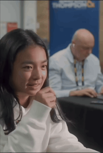 A Girl Sits At A Table With A Man Behind Her And A Sign That Says Troop Room On It GIF