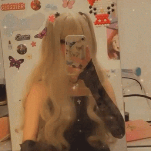 A Girl Taking A Selfie In Front Of A Mirror With Stickers On The Wall That Say Glossier GIF