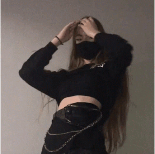 A Girl Wearing A Mask And A Black Crop Top Is Standing In Front Of A Wall . GIF