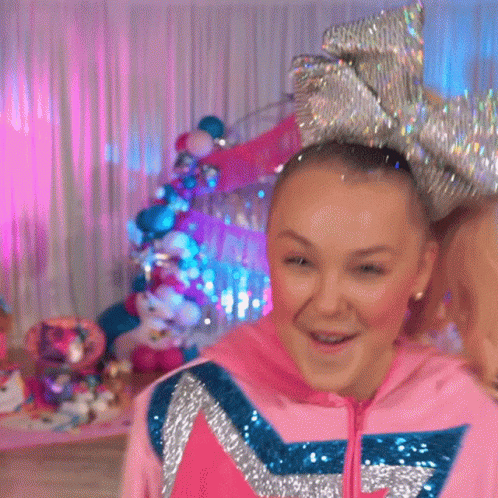 A Girl Wearing A Pink Hoodie And A Large Bow In Her Hair Is Standing In A Room . GIF