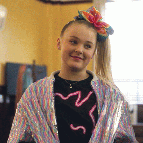 A Girl Wearing A Rainbow Jacket And A Bow In Her Hair Smiles GIF