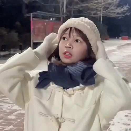 A Girl Wearing A White Hat And Scarf Holds Her Hands To Her Head GIF