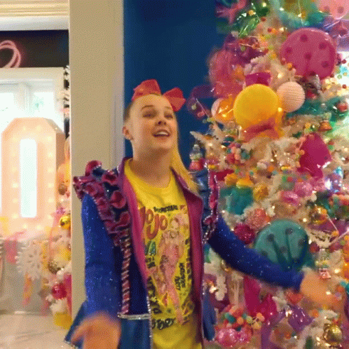 A Girl Wearing A Yellow Shirt That Says ' Jojo ' On It Is Dancing In Front Of A Christmas Tree GIF