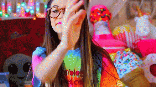 A Girl Wearing Glasses And A Tie Dye Shirt Is Making A Peace Sign . GIF