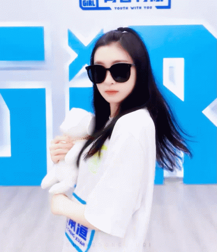 A Girl Wearing Sunglasses Is Holding A Stuffed Animal In Front Of A Sign That Says Girl Youth With You GIF