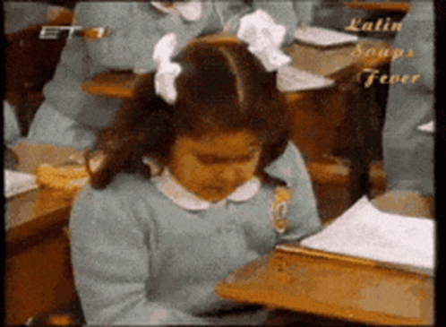 A Girl With A Bow In Her Hair Sits At A Desk In A Classroom With Latin Soups Fever Written On The Bottom GIF