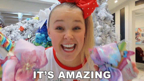 A Girl With A Red Bow On Her Head Says It 'S Amazing In Front Of A Christmas Tree GIF