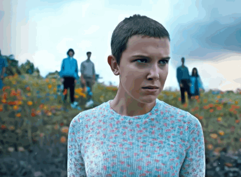 A Girl With A Very Short Haircut Is Standing In A Field Of Flowers GIF