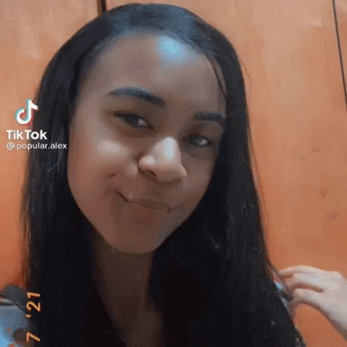 A Girl With Long Black Hair Is Smiling And Looking At The Camera While Holding Her Hair . GIF