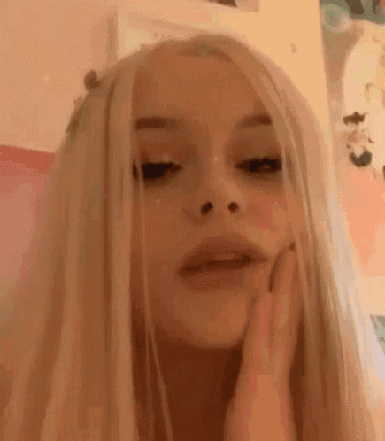 A Girl With Long Blonde Hair Is Making A Funny Face While Touching Her Face . GIF