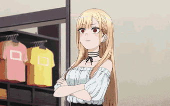 A Girl With Long Blonde Hair Is Standing With Her Arms Crossed In Front Of A Clothes Rack . GIF
