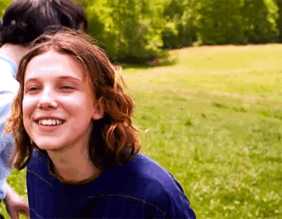A Girl With Long Hair Is Smiling While Standing In A Field . GIF