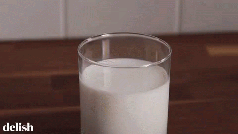 A Glass Of Milk Is Sitting On A Wooden Table With The Word Delish On The Bottom . GIF