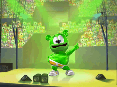 A Green Gummy Bear Is Standing On A Stage In Front Of A Crowd GIF