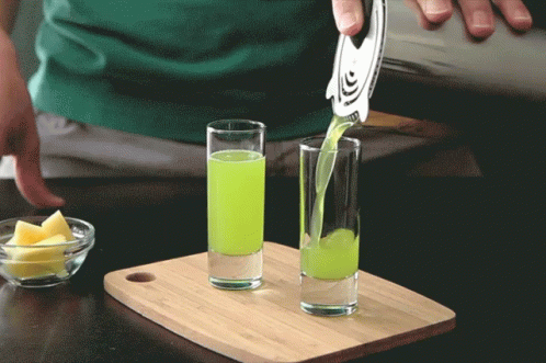 A Green Liquid Is Being Poured Into Two Shot Glasses On A Cutting Board GIF