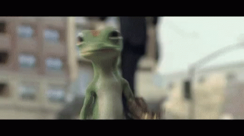 A Green Lizard Is Standing In Front Of A Building And Looking At The Camera GIF