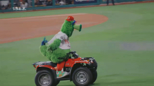 A Green Mascot Is Riding A Four Wheeler On A Baseball Field Sponsored By Phillies GIF