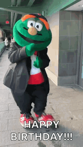 A Green Mascot Is Standing On A Sidewalk And Says Happy Birthday ! GIF