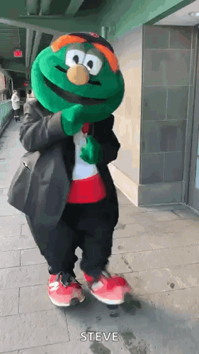 A Green Mascot Is Standing On A Sidewalk Wearing A Suit And A Hat . GIF