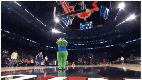 A Green Mascot Stands On A Basketball Court In Front Of A Scoreboard That Says Coca Cola GIF