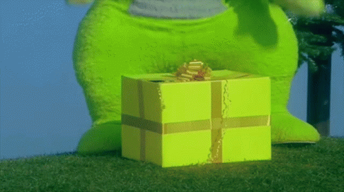 A Green Stuffed Animal Standing Next To A Yellow Gift Box With A Gold Bow GIF