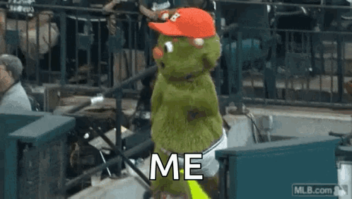 A Green Stuffed Animal Wearing A Red Hat Is Standing In A Stadium . GIF