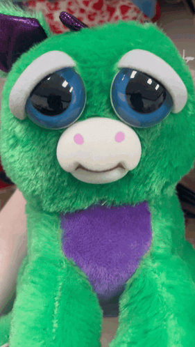 A Green Stuffed Animal With Blue Eyes And A Purple Heart On Its Chest GIF