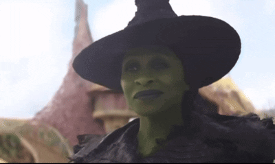 A Green Witch With A Black Hat Is Standing In Front Of A Building GIF